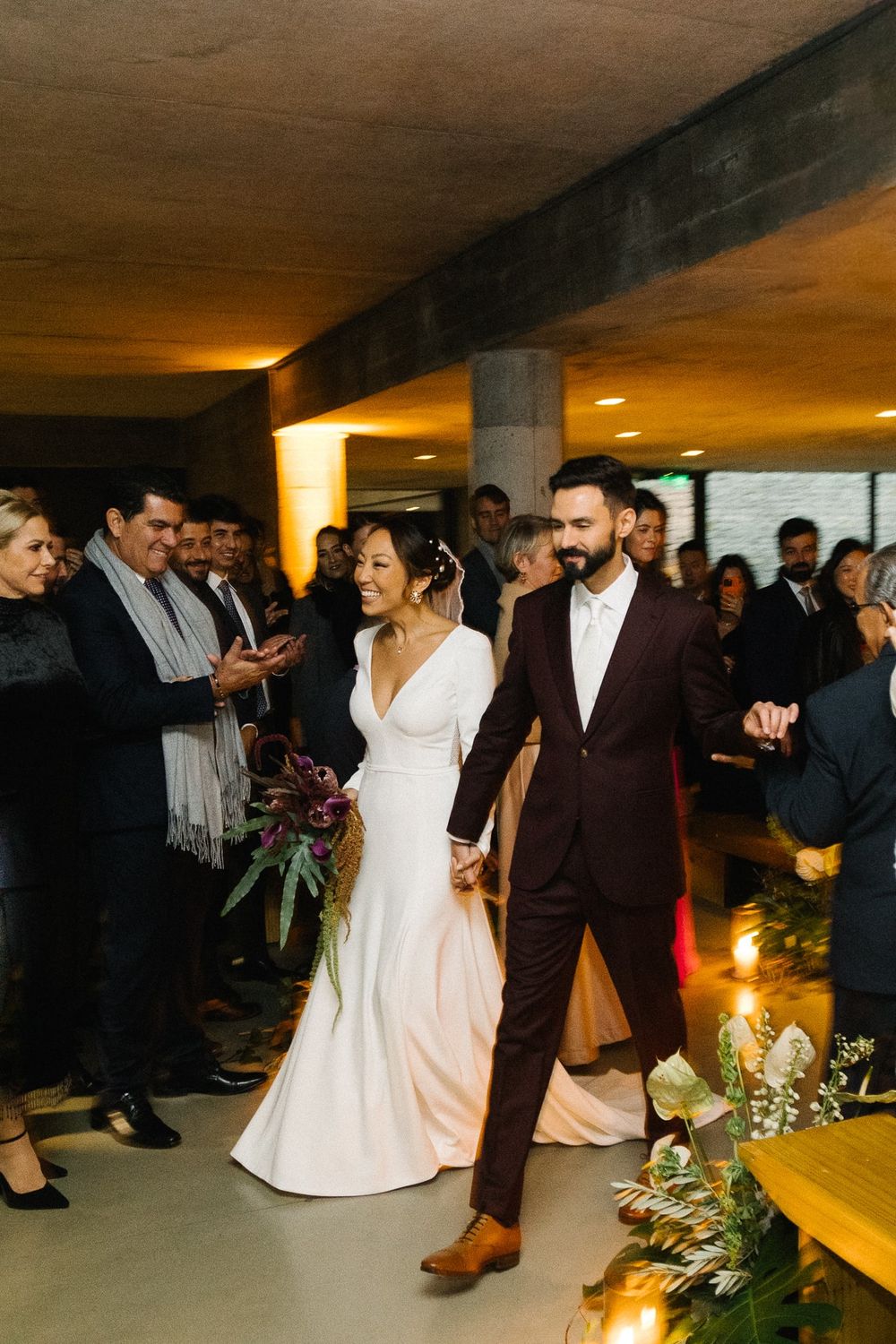 Newlyweds walking down aisle surrounded by guests in industrial venue space.