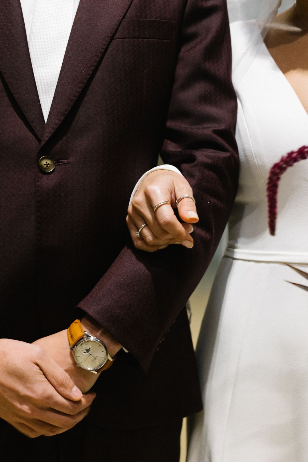 Detail shot of groom's burgundy suit jacket and watch while holding hands.