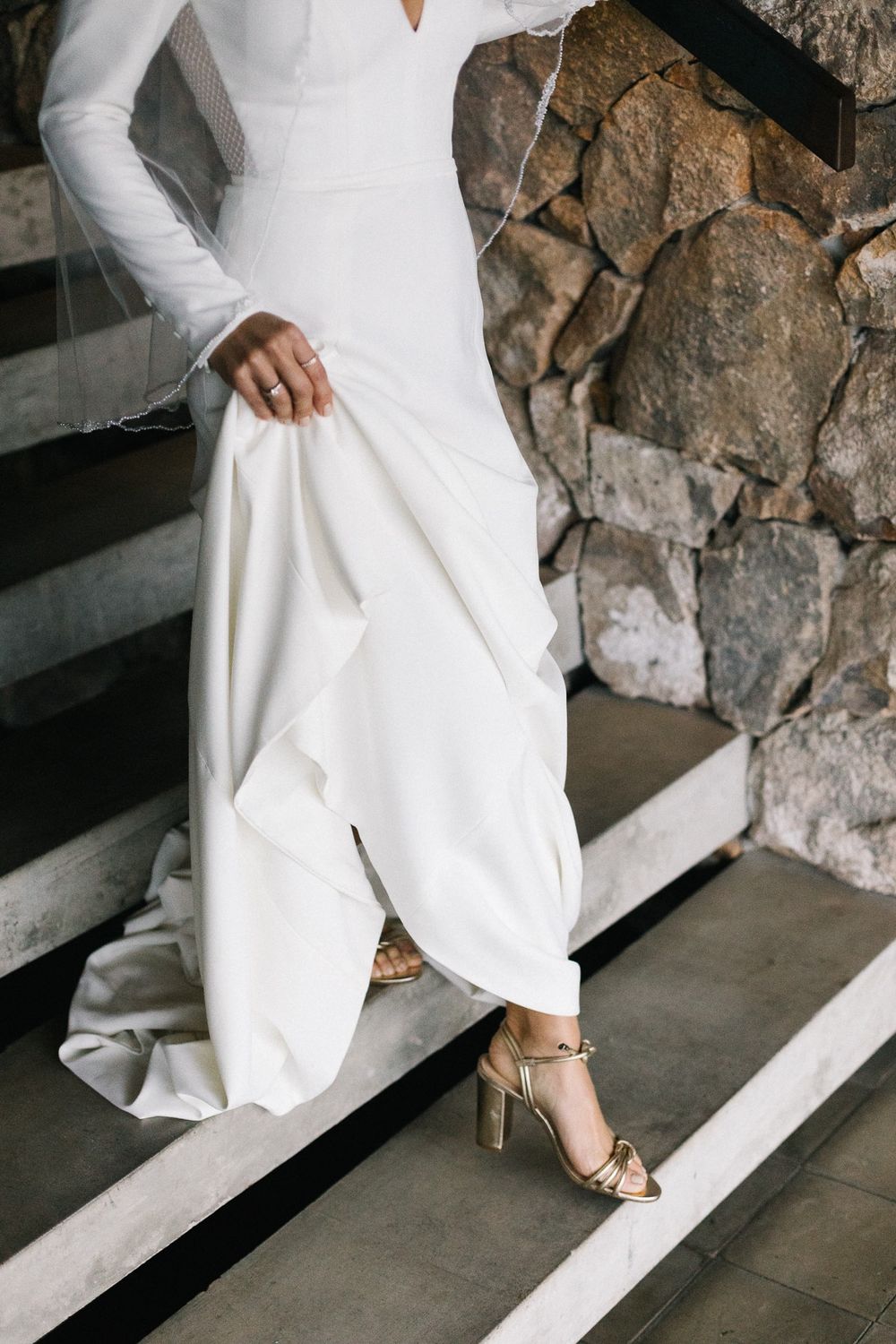 Detail shot of a sleek white wedding dress and gold heels on concrete steps.