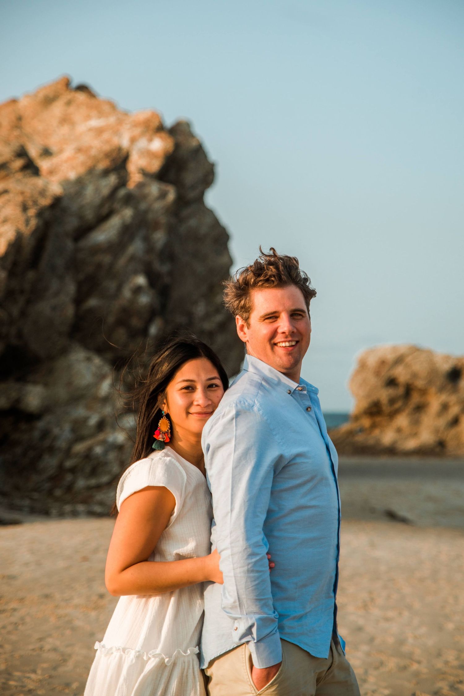 A couple poses together on a beach with dramatic rocky cliffs in the background.