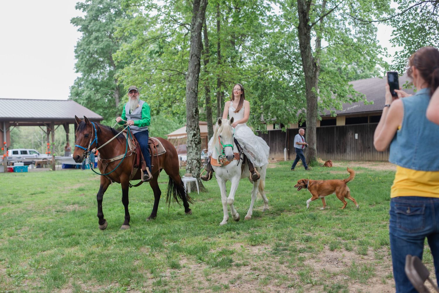 Horses and riders on a grassy field with a dog running alongside.
