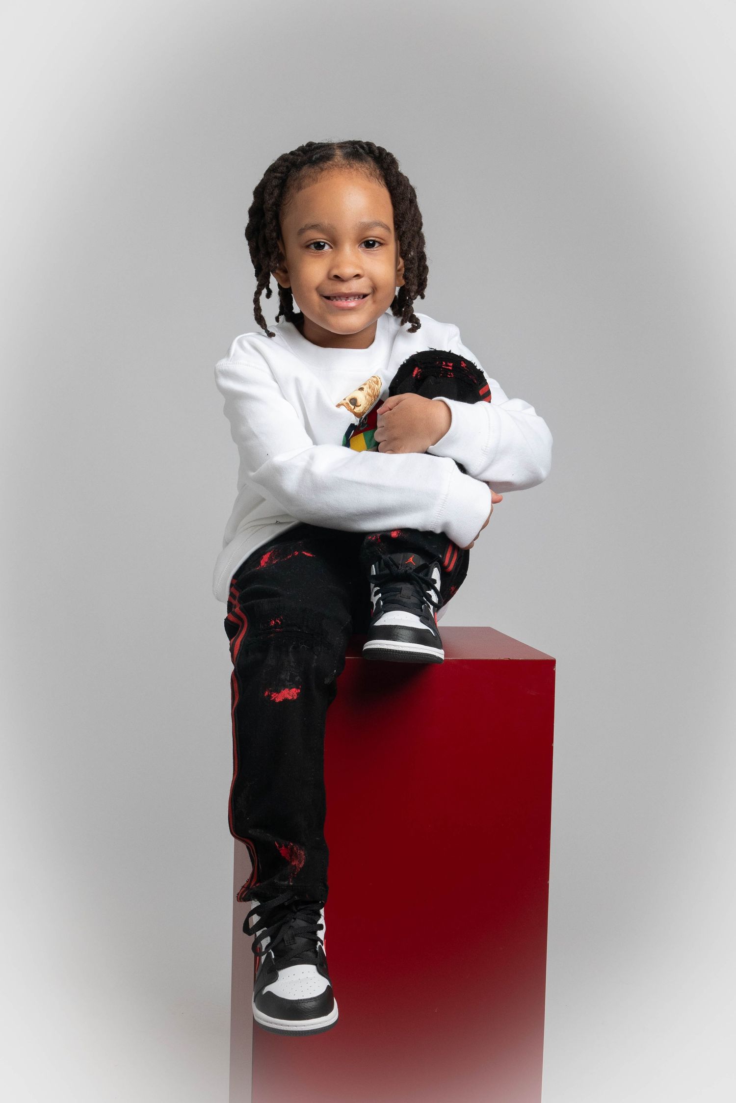 Child in white sweater and patterned pants posing on red bench against light background.