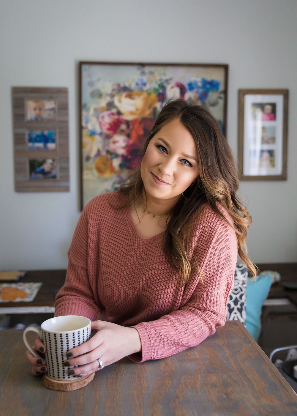Woman in pink sweater enjoys coffee at wooden table with colorful artwork displayed on wall behind her.
