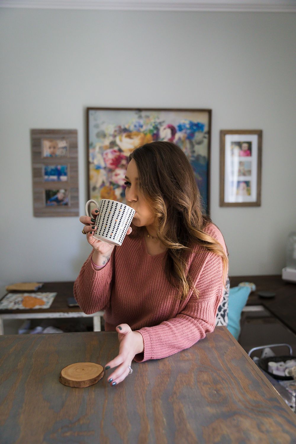 Person in cozy pink sweater sips from striped mug at wooden table with artwork in background.