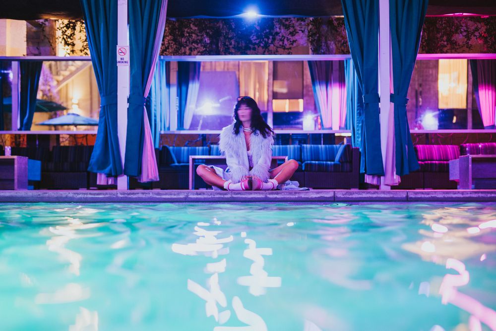 JJ Wilde sits on a pools edge in Arizona with a blurred face in a blue fuzzy jacket and lingerie