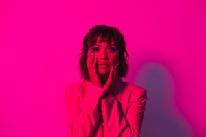 JJ WILDE has both hands cradling her face, looking into the camera, the photo is saturated neon pink.