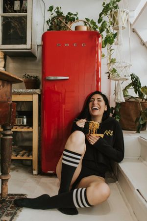 Marie Nicola sits on the floor eating noodles with chopsticks in front of a red SMEG fridge wearing knee high socks.