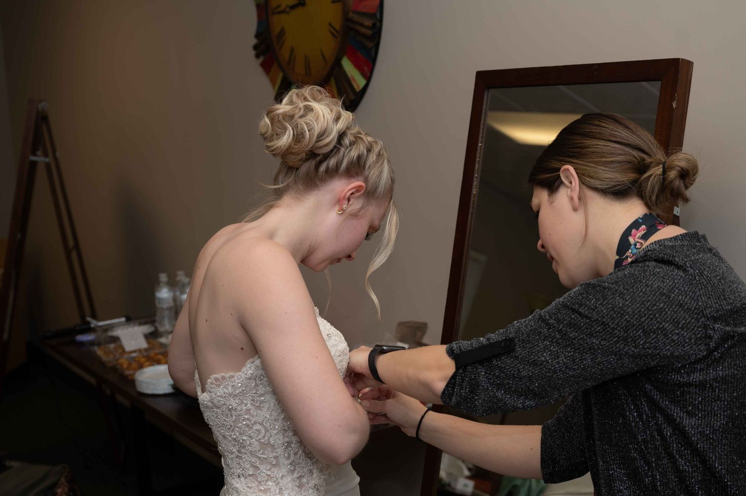 A person in a wedding dress gets help with final preparations in a dressing room.
