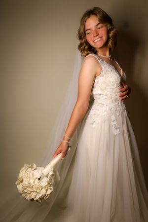 A bride in a flowing white lace gown holds a white bouquet while posing against a neutral background.