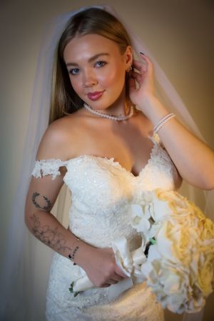 Bride with tattoo holds white rose bouquet in classic strapless wedding gown.