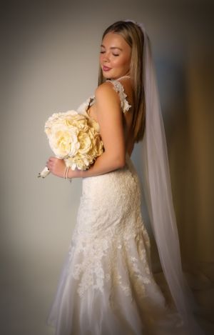 A bride in a fitted lace gown holds a cream bouquet while posing against a neutral background.