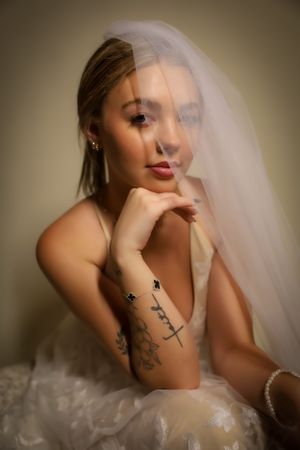 A bride with visible tattoos poses thoughtfully in her wedding gown and veil.