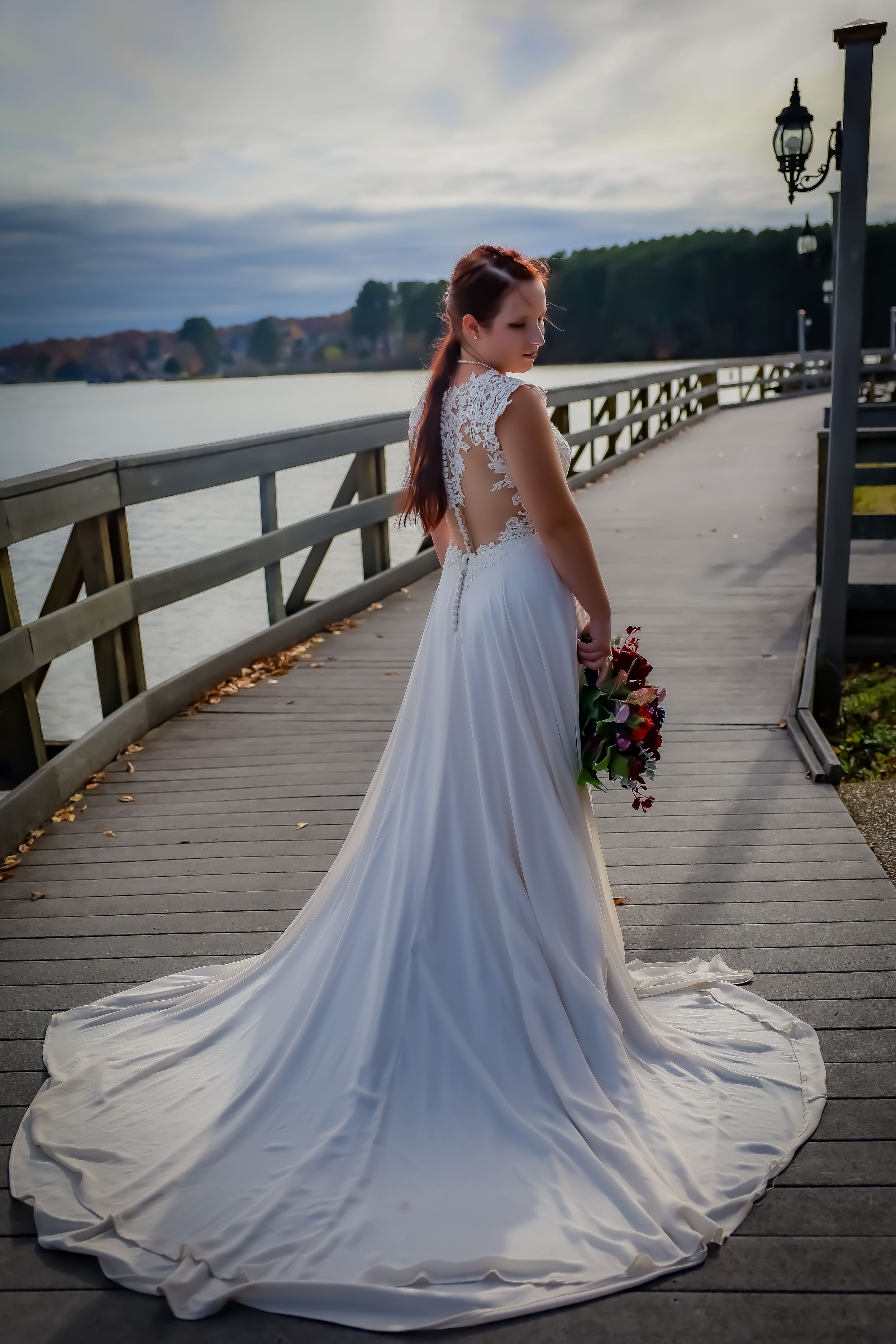 Bride in flowing white gown poses on wooden boardwalk by waterfront.