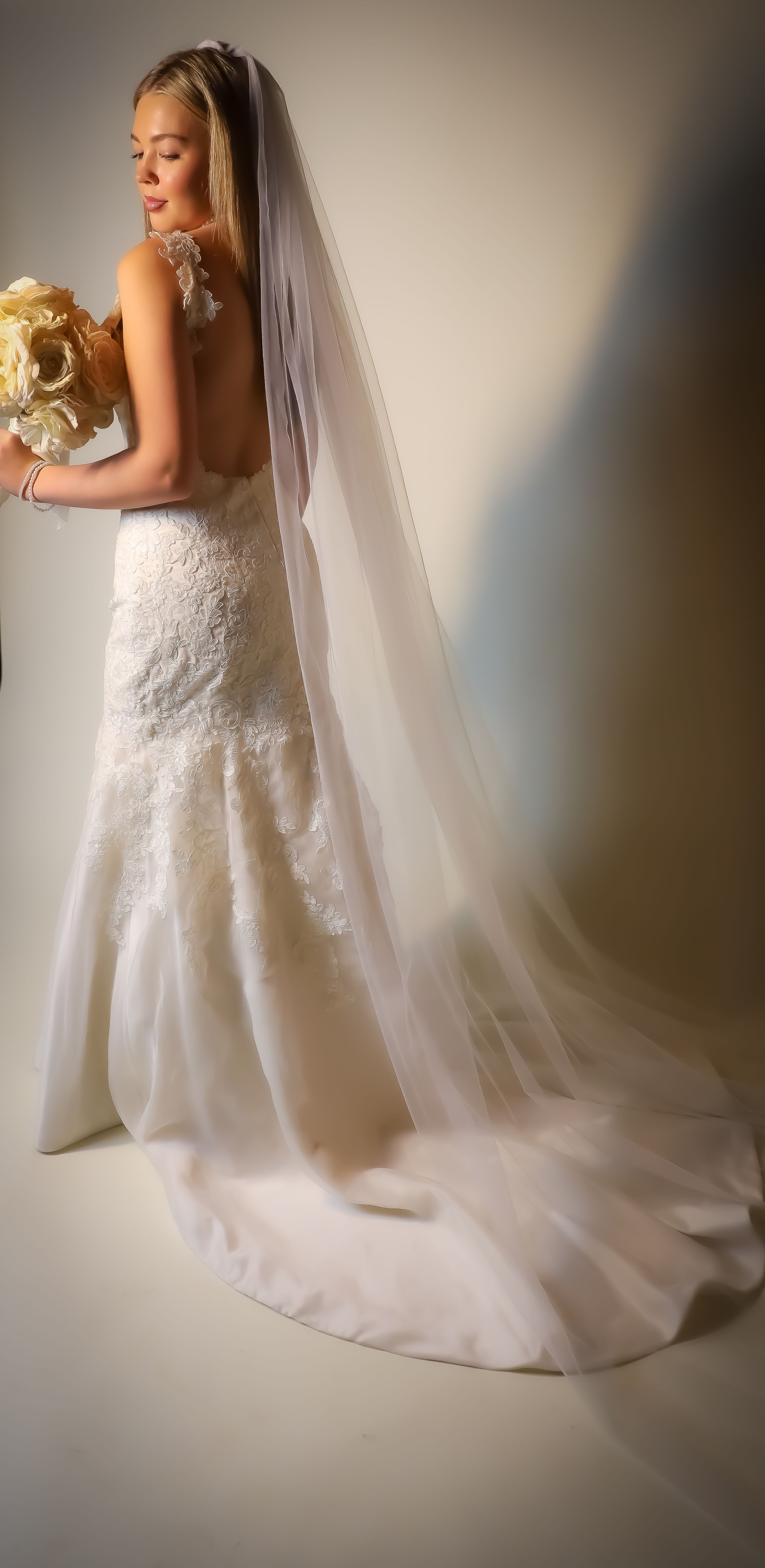 A bride in a lace wedding gown with long train holds a cream bouquet against a neutral backdrop.