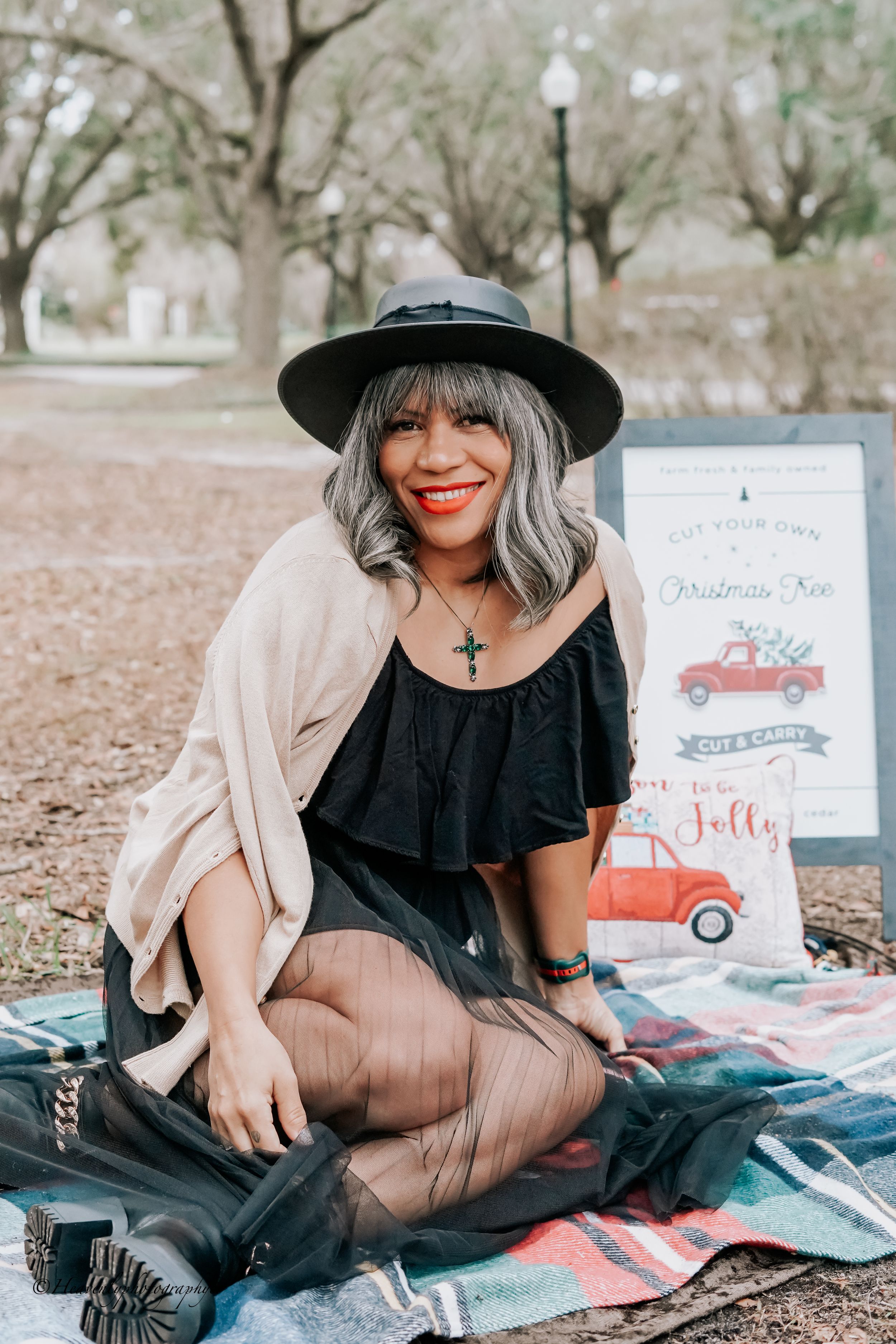 Woman in black hat and beige cardigan poses with holiday-themed artwork on picnic blanket.