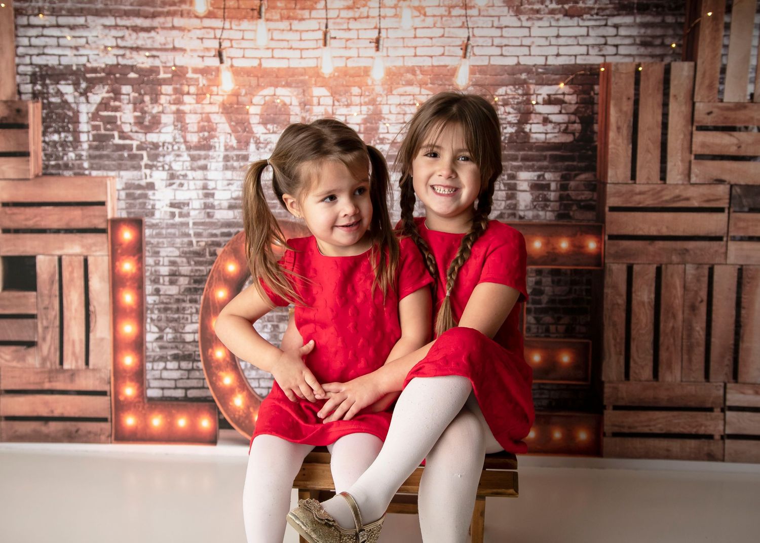 Two children in red dresses pose together against brick wall with illuminated LOVE letters.