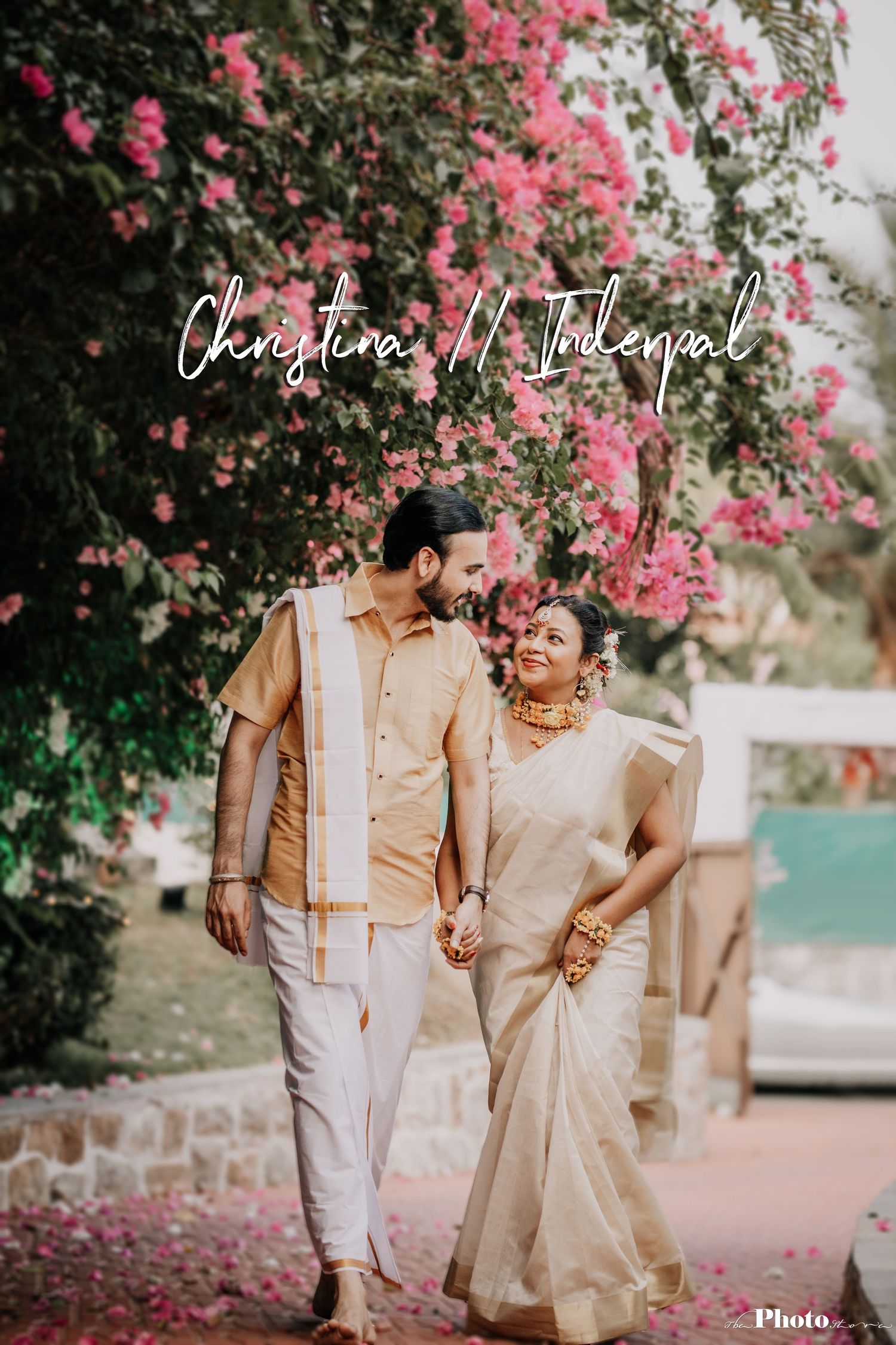 Couple walks hand in hand under blooming pink bougainvillea in traditional cream-colored attire.