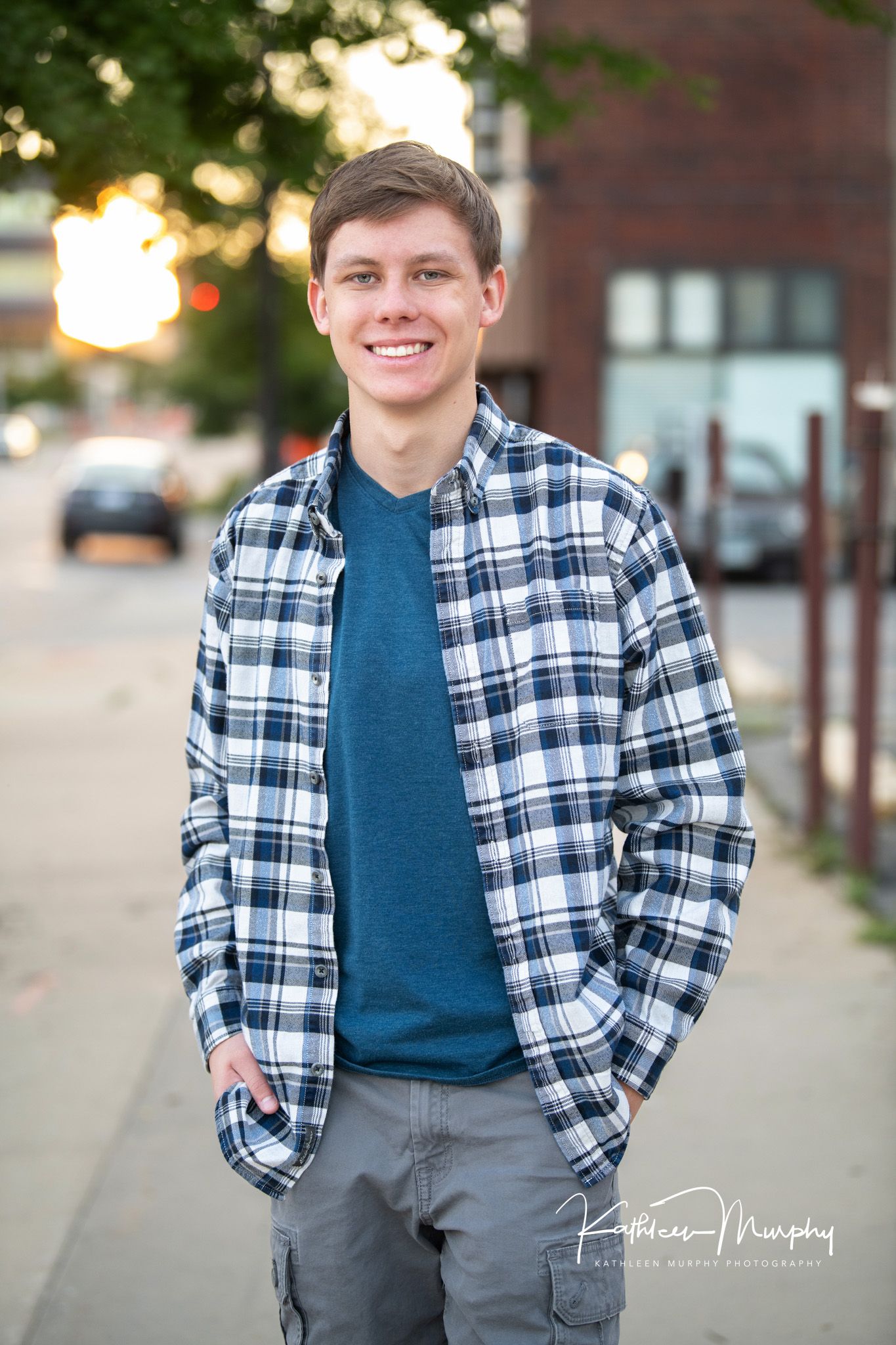 Person in plaid shirt and blue t-shirt standing outdoors at sunset with brick building in background.