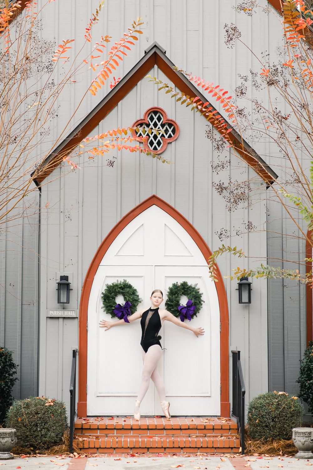 Ballerina poses for photos in fourth position en pointe wearing a leotard against white chapel doors with purple wreaths in Bluffton, SC.