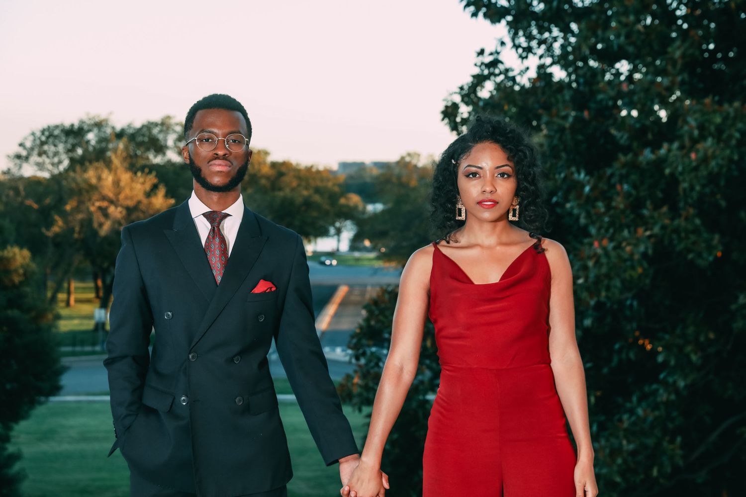 A stylish couple holds hands outdoors, man in black suit, woman in red dress, with trees behind them.