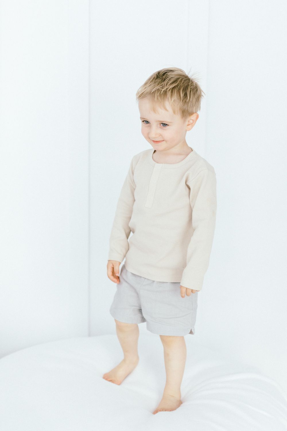 Toddler in beige sweater and gray shorts standing barefoot on white surface.