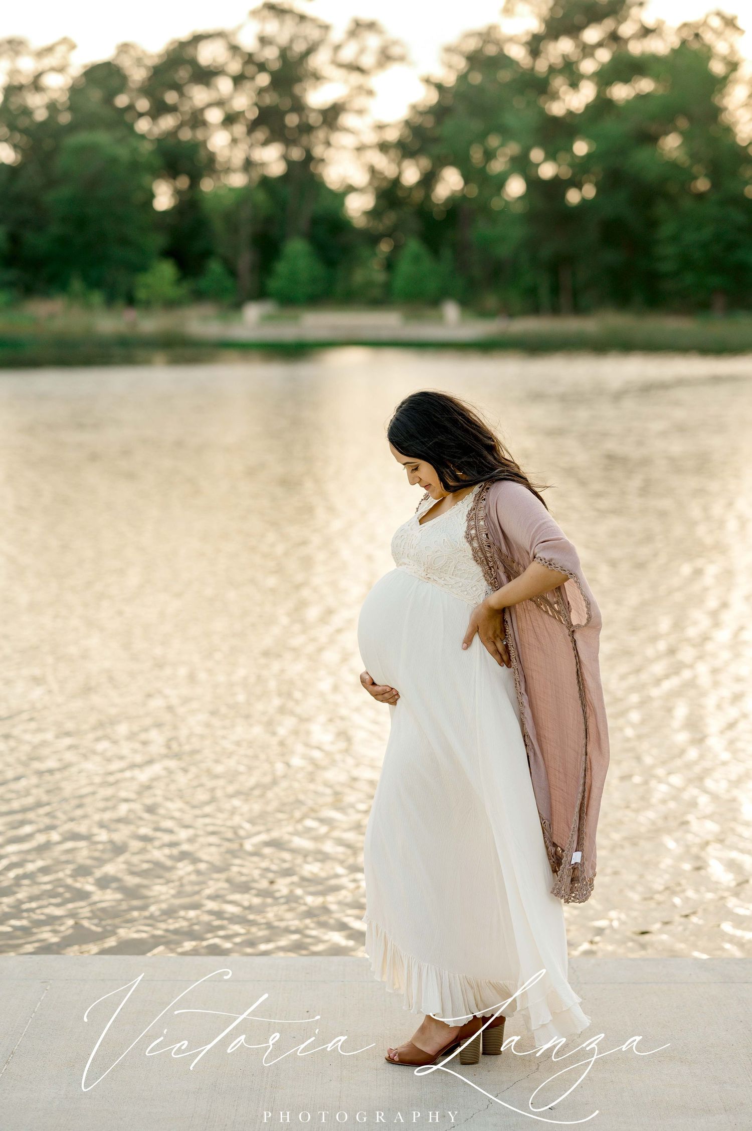 pregnant woman wearing white lace dress posing for a maternity photo session standing behind a lake at sunset