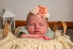 Sleeping newborn baby girl wearing a pink floral headband, resting in a woven basket with cream knit blanket.