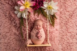 Newborn baby swaddled in pink on wooden swing surrounded by colorful flowers.