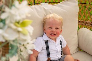 Adorable blonde toddler boy in white shirt with suspenders smiling on a cozy chair.