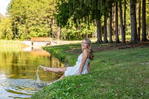 Young woman in white dress sitting by a peaceful pond splashing water on a sunny day.