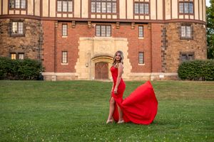 Woman in flowing red gown poses on green lawn before historic brick building at golden hour.
