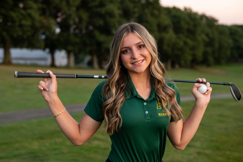 Young woman in green golf polo holds club behind shoulders on golf course at dusk.