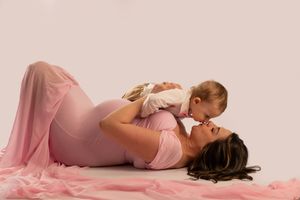 Pregnant woman in pink dress lying down while playing with her smiling baby girl.