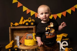 Baby boy in construction themed shirt celebrating first birthday with yellow cake smash and toy trucks.