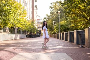 Young woman in a white dress posing on a brick walkway lined with trees in an urban setting.