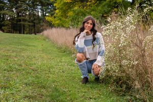 Young woman crouching near wildflowers in a green meadow, wearing ripped jeans and a patchwork sweater.