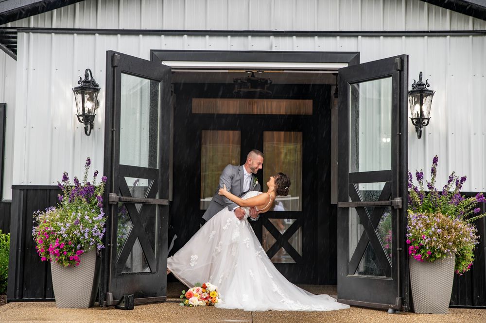 Groom dips bride in white gown at elegant black and white barn wedding venue entrance with floral decor.
