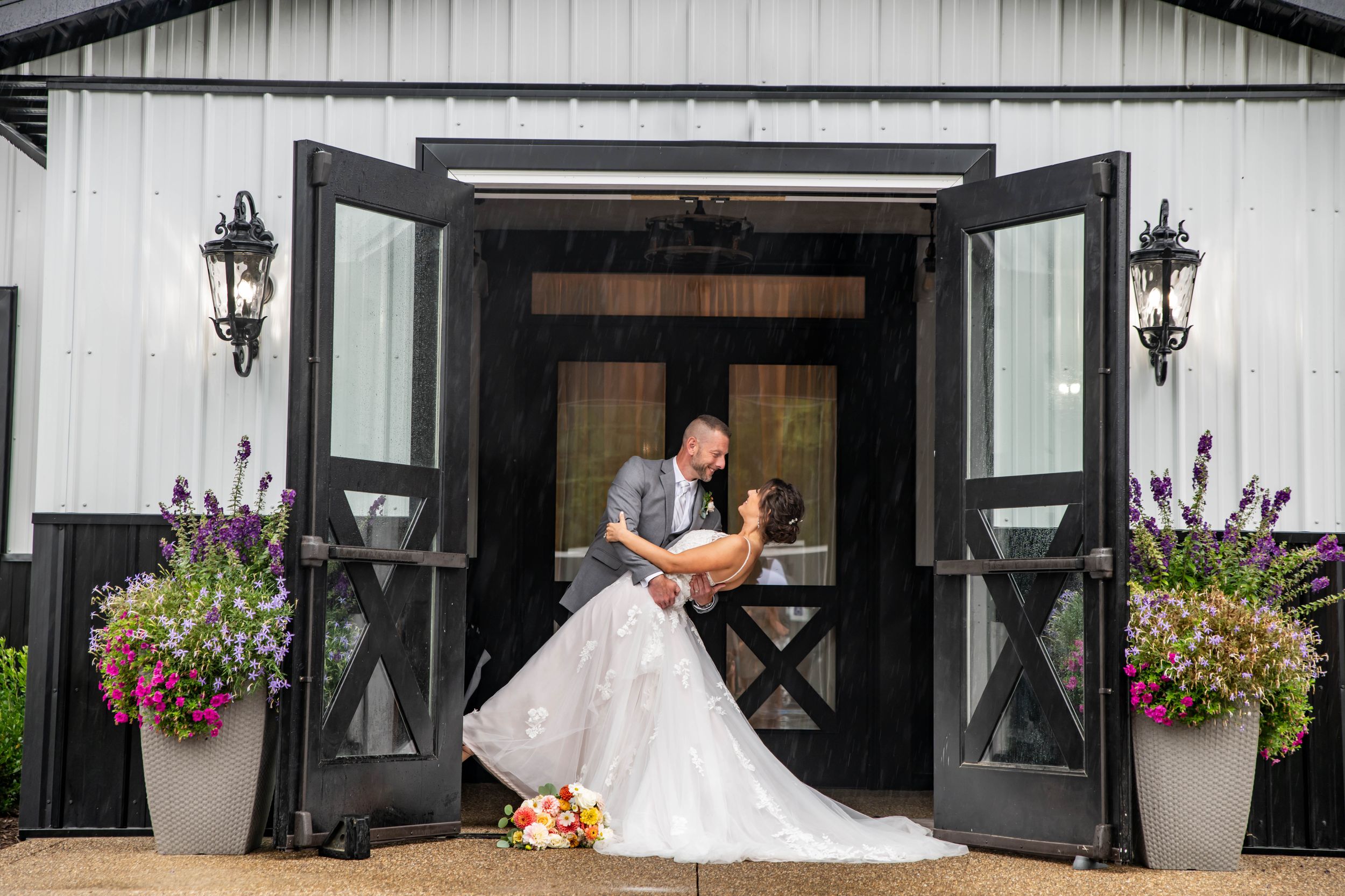 Groom dips bride in white gown at elegant black and white barn wedding venue entrance with floral decor.