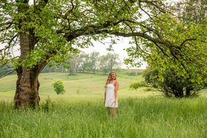 Young woman in white dress standing in lush green field beneath arching oak tree.
