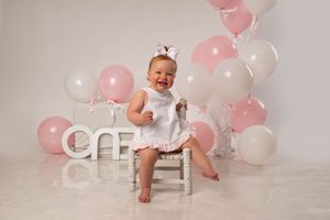 Baby girl in white dress laughing at first birthday cake smash with pink and white balloons.
