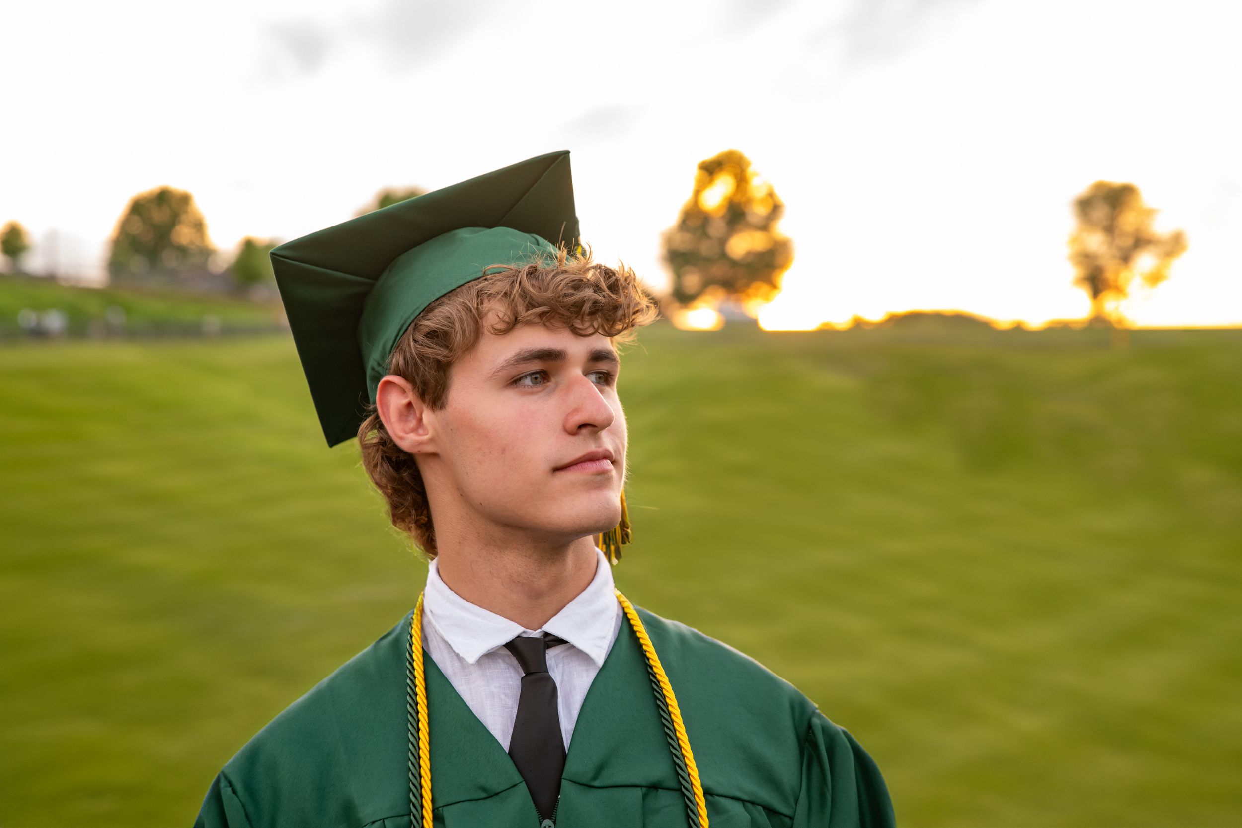 Young man in green graduation cap and gown with black tie poses outdoors on a green hillside at sunset.