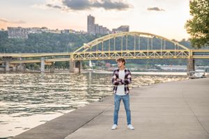 A young man stands by the riverside with Pittsburgh's iconic yellow bridge behind him.