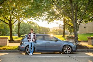 Young man leaning on blue sedan under golden autumn trees at sunset on tree-lined street.