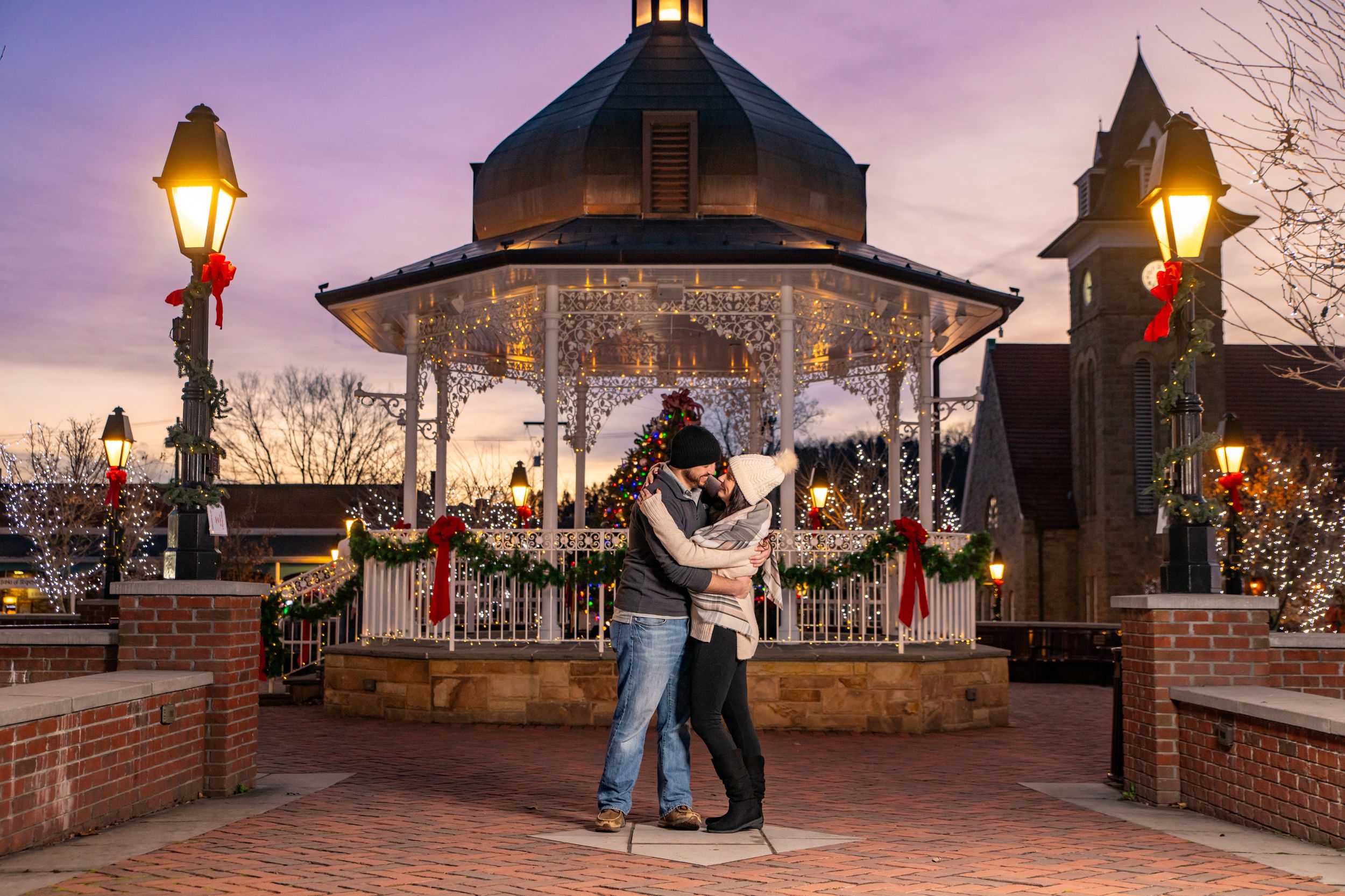 Couple kissing near a decorated gazebo with Christmas lights at twilight in a festive town square.
