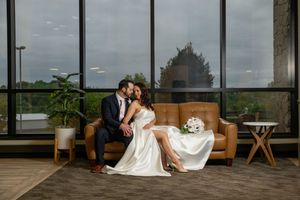 Bride and groom sharing a kiss on a leather sofa in front of large panoramic windows.