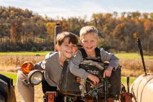 Two smiling young boys pose together on a vintage tractor in an autumn farm field.
