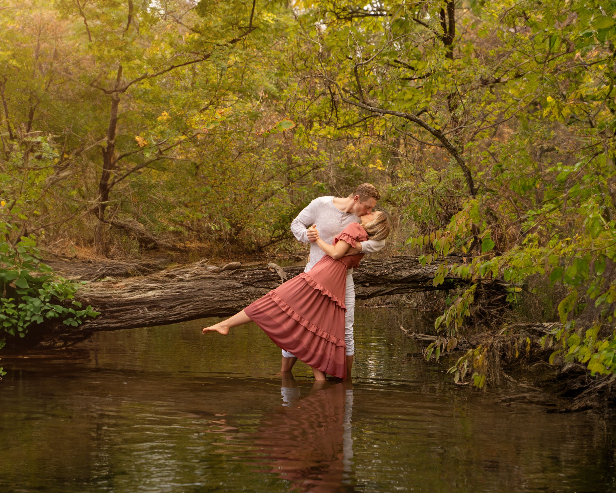 Couple sharing a romantic embrace while standing in a shallow creek surrounded by lush autumn foliage.