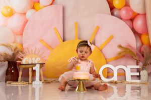 Baby girl in pink outfit touching first birthday cake with sunshine themed balloon backdrop.