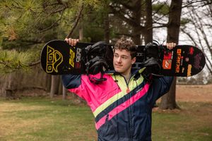 Young man holding a snowboard on his shoulders outdoors wearing a colorful pink and yellow winter jacket.