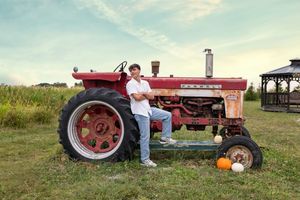 Young man leaning against vintage red Farmall tractor in a farm field with pumpkins nearby.